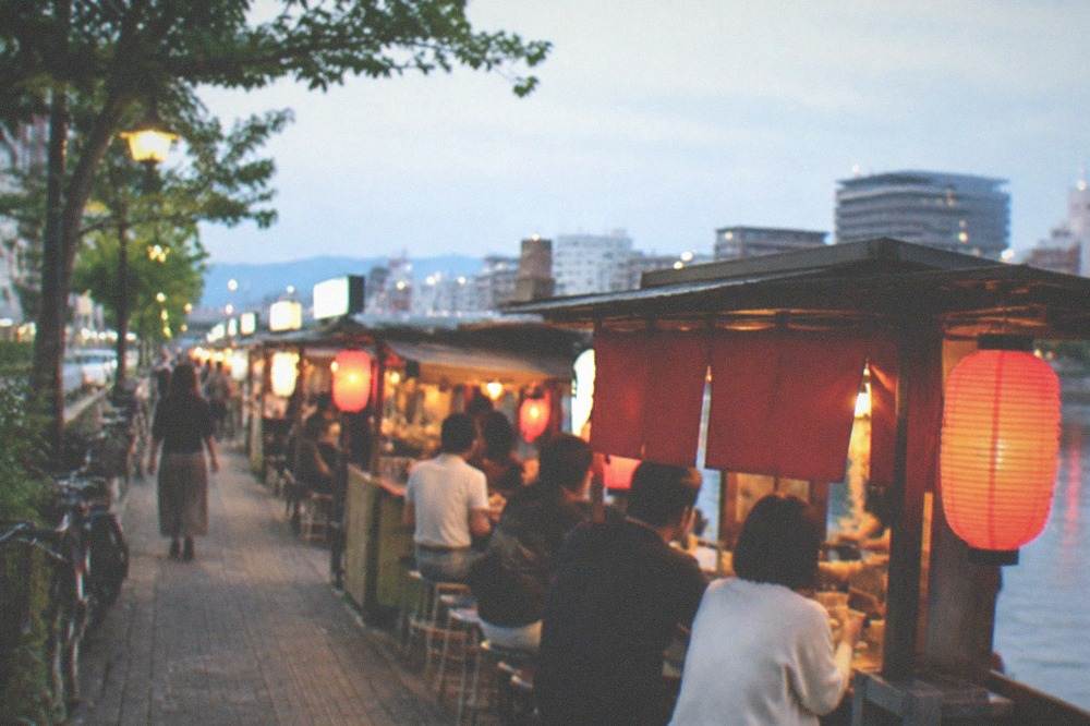 A row of lit food stalls along a river in Fukuoka at night with customers seated at counters