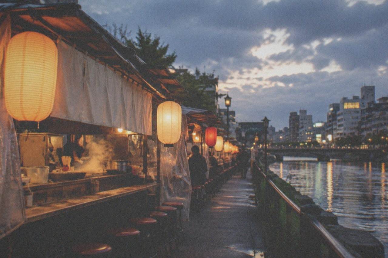 Food stalls with lanterns glowing along a riverside street in the evening