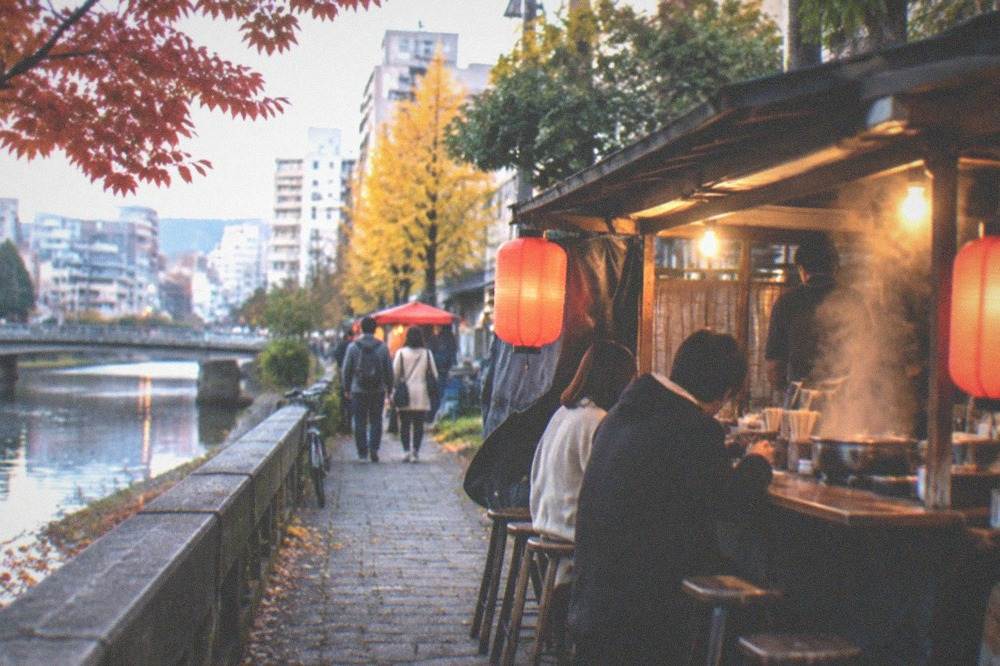 A glowing food stall beside a river at night with steam rising from a hot pot
