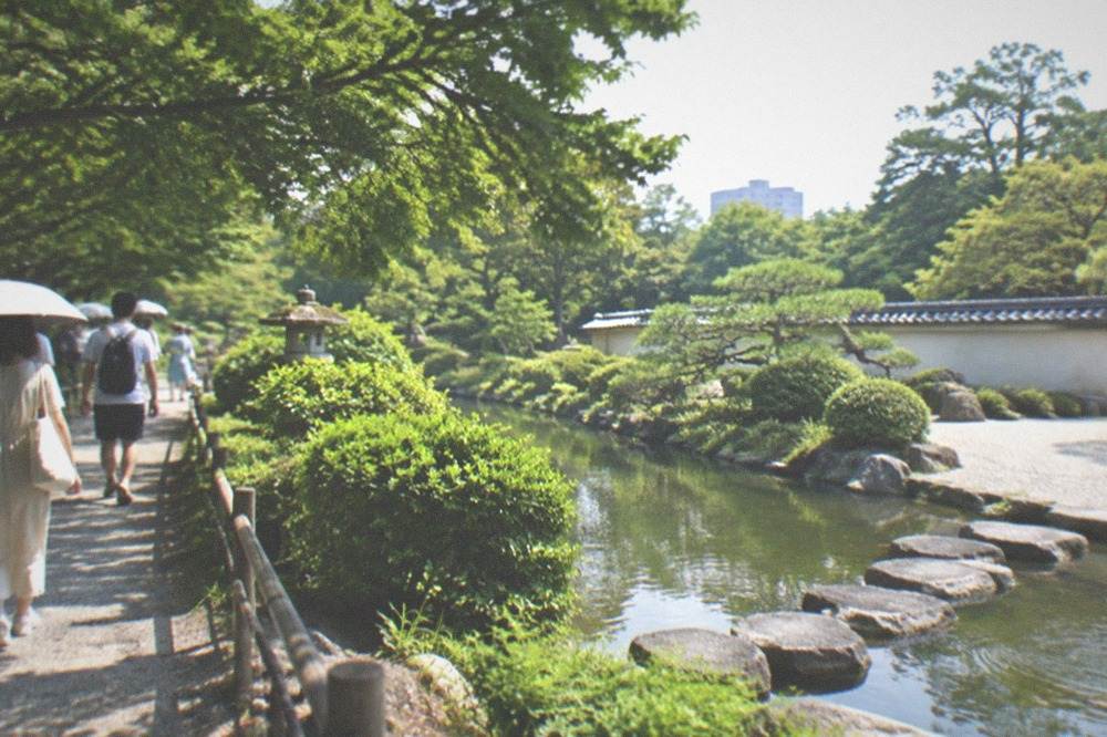 Visitors walking a shaded path beside the pond in the Ohori Park Japanese Garden