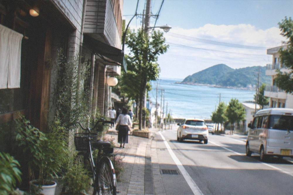 A coastal road with green hills meeting a calm blue sea under scattered clouds