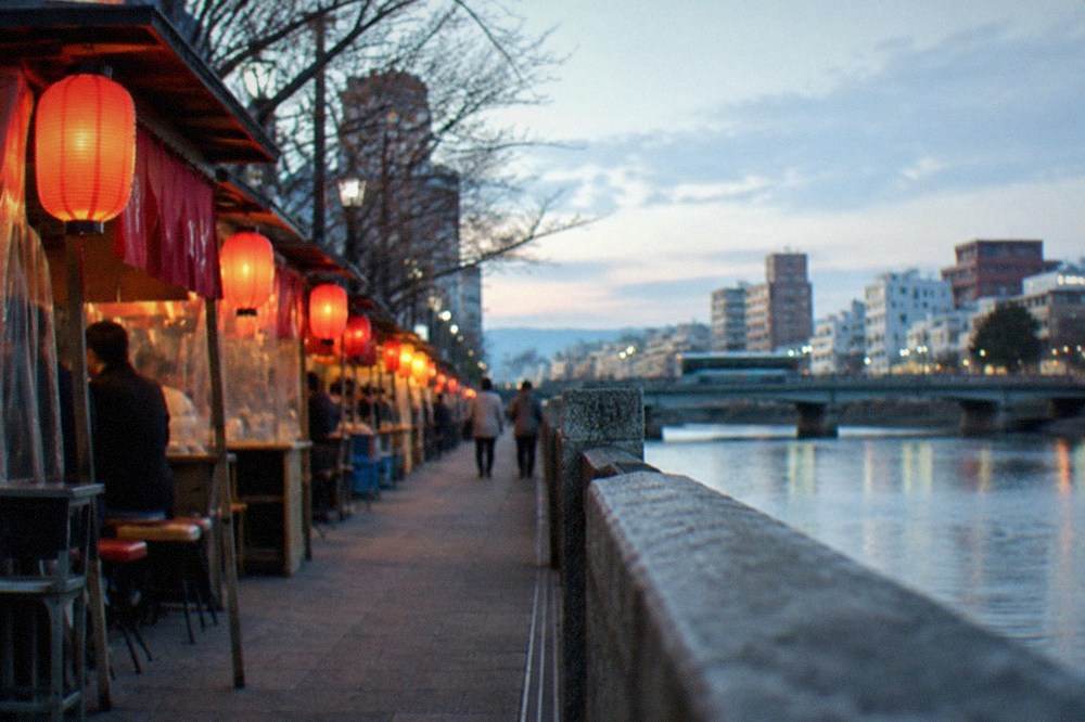 A row of food stalls with red lanterns glowing along a riverside walkway at night