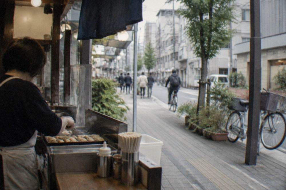 A cook preparing food at a small counter stall with a hand-written price menu displayed