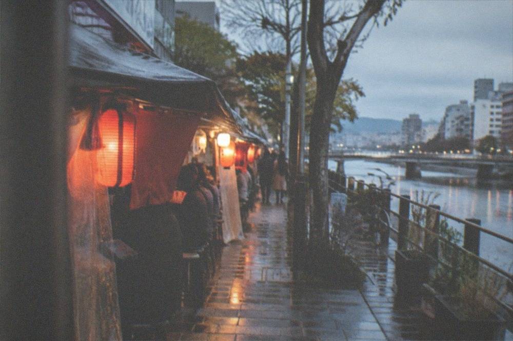 A row of food stalls along a riverside street with rain slicked awnings and warm lantern light