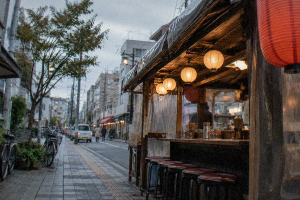 A narrow lantern-lit food stall with counter seating beside a city street