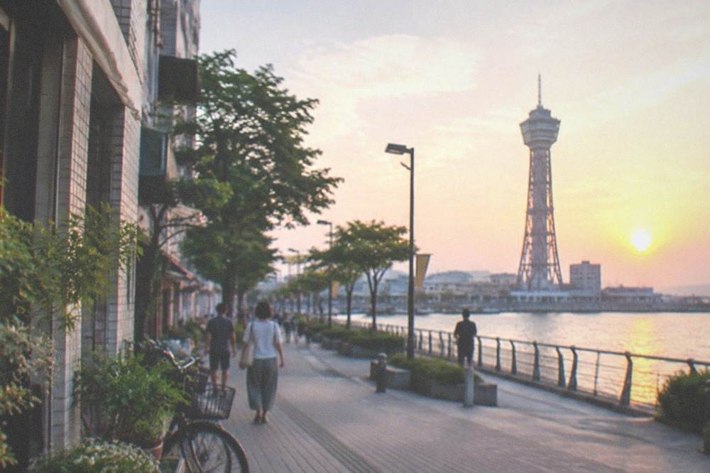 A tall lattice tower standing above a waterfront promenade at sunset