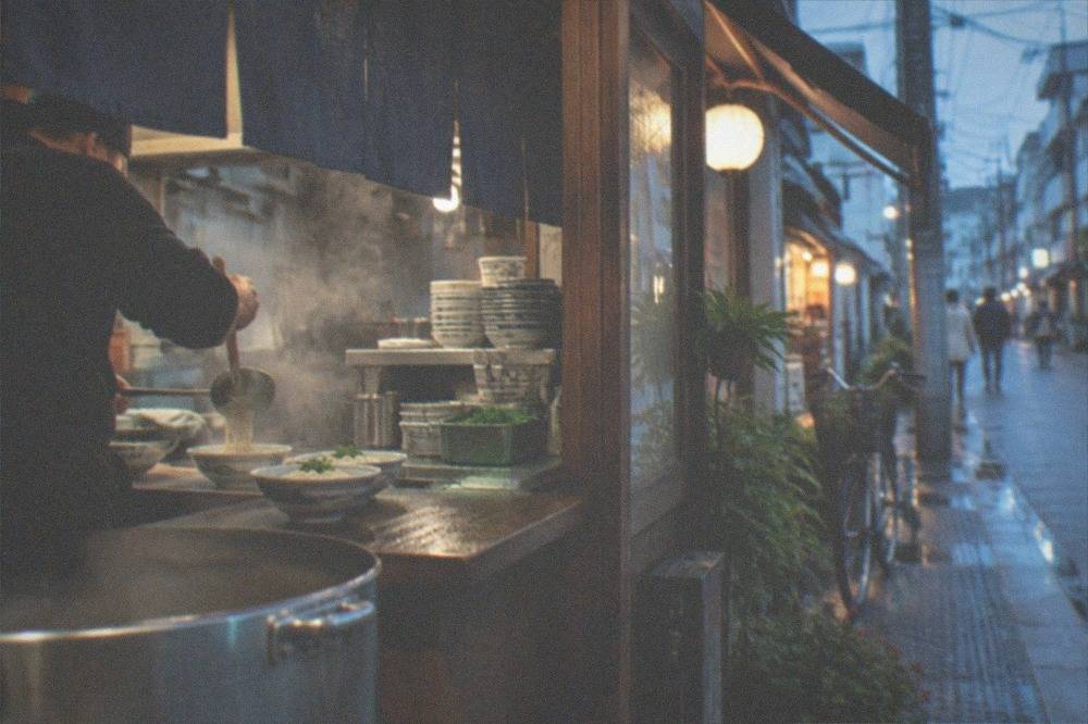 A small udon shop counter with a steaming pot and a cook ladling noodles into bowls