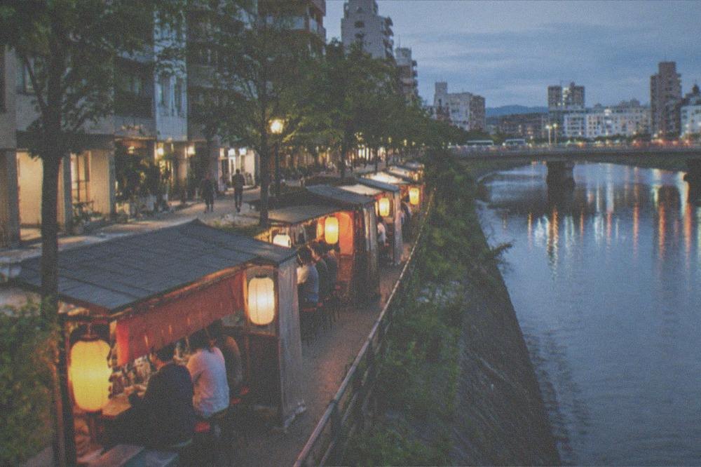 A row of lantern-lit food stalls along a river at night with customers seated at counters