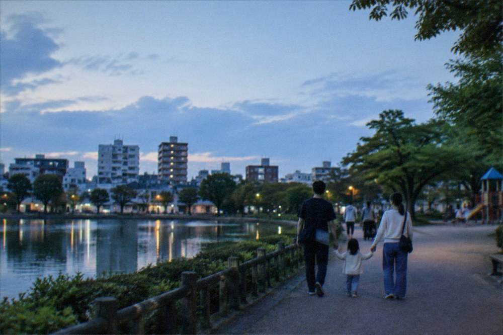 Families walking a wide park path beside a pond with trees and a playground in the distance