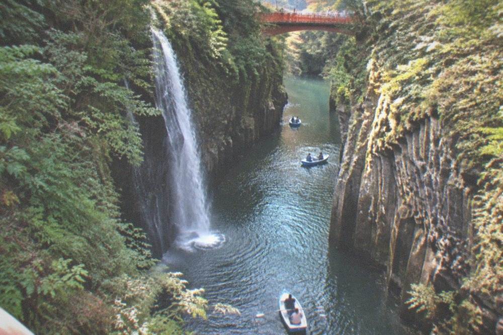 Red footbridge over a wooded gorge with boats on green water below at Takachiho