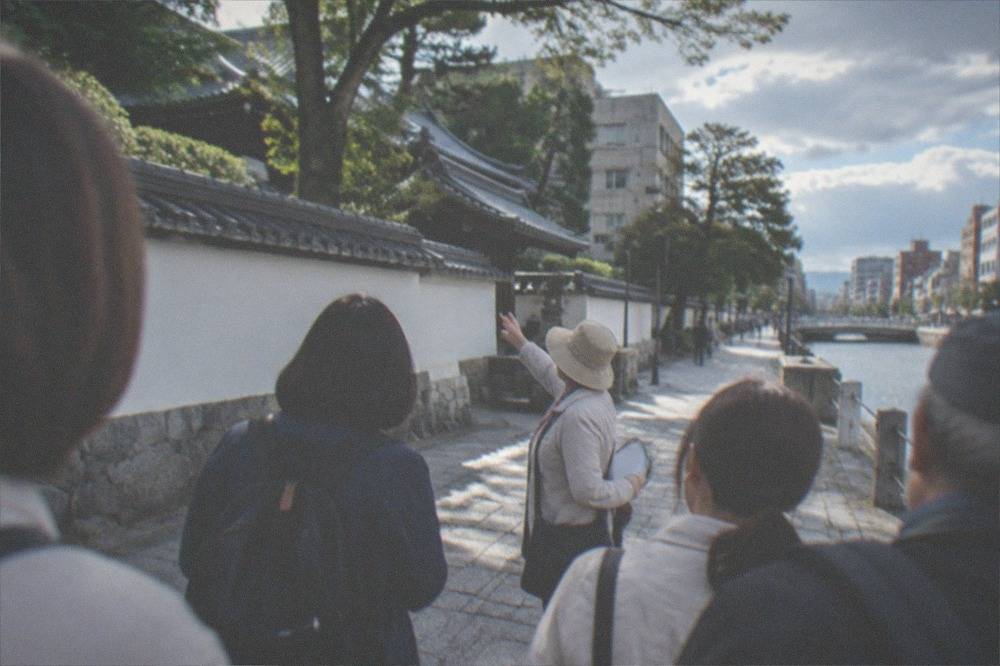 A guide pointing out buildings along a canal-side promenade while a small group listens