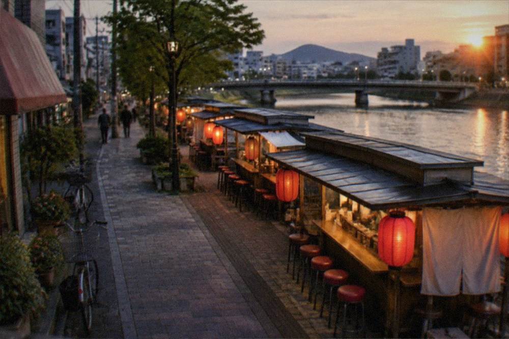 A row of small lit food stalls with red lanterns lined along a river at dusk, counter stools empty.