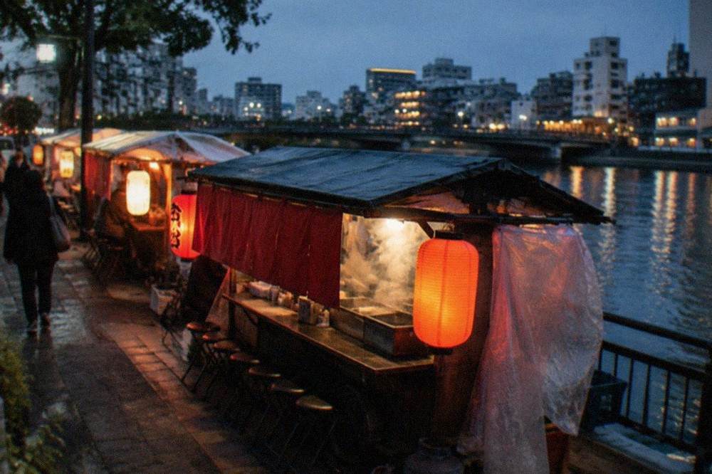 Narrow yatai stall with red lanterns and steam rising from a cooking counter beside a city street