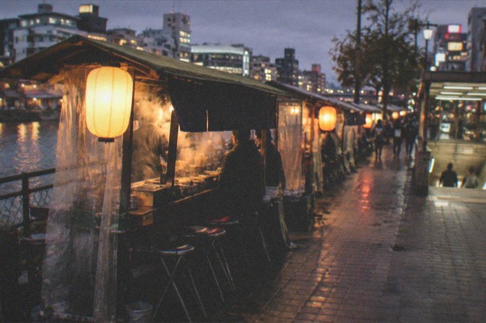 Illuminated city street at night with a subway station entrance and distant lanterns