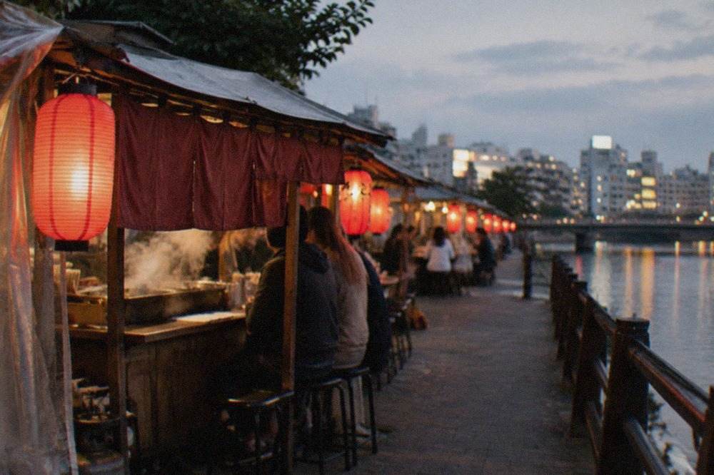 Small lit food stalls with red lanterns lined along a riverside walkway at night