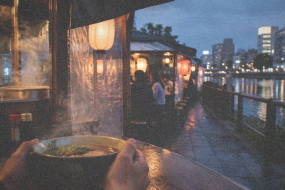 Close view of hands holding a bowl of noodles at a narrow wooden food stall counter at night