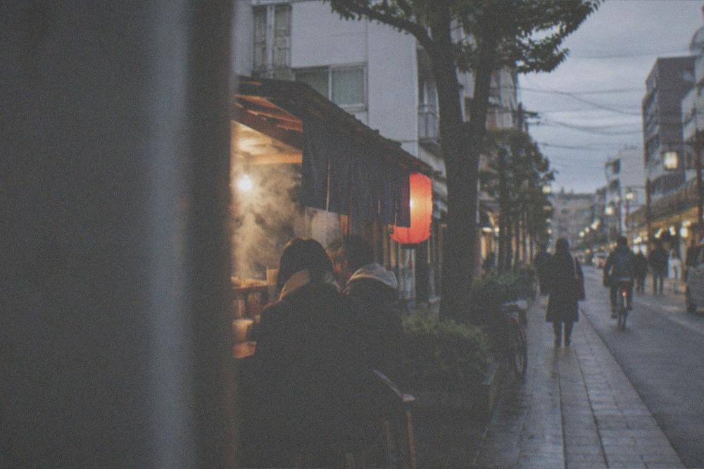 Food stall cart with steam and warm lights on a cool evening street with a few seated customers