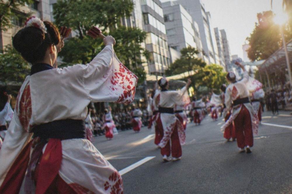 Costumed parade participants dancing along a wide city avenue lined with tall buildings