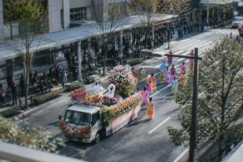 A decorated flower-covered parade car surrounded by marchers in bright costumes on a city street