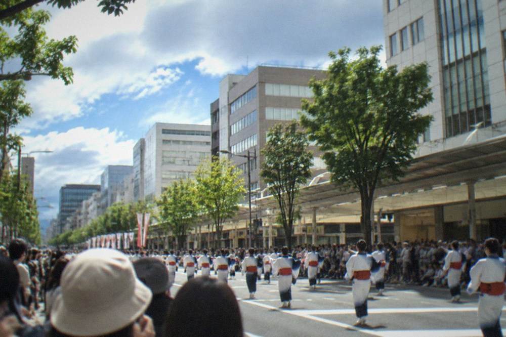 Crowds of spectators lining a broad avenue while a daytime parade passes between tall buildings