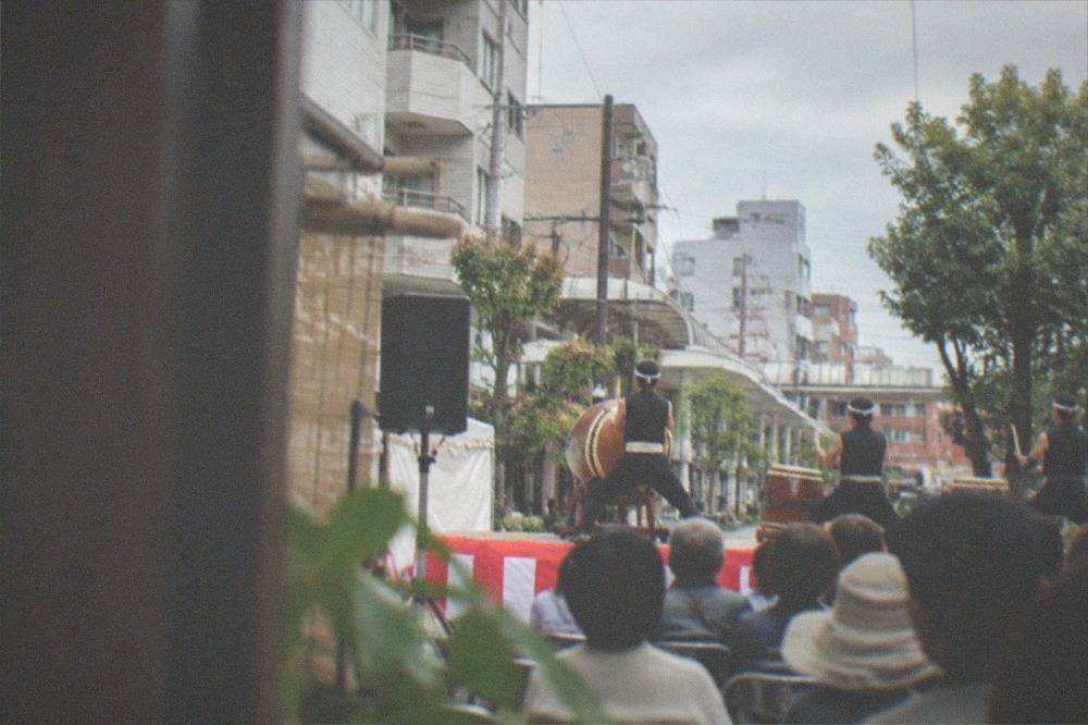 Drummers performing on an outdoor stage in front of a seated street crowd