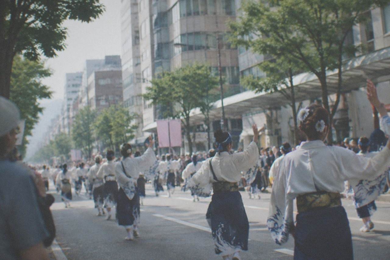 Costumed parade participants dancing along a wide city avenue lined with tall buildings