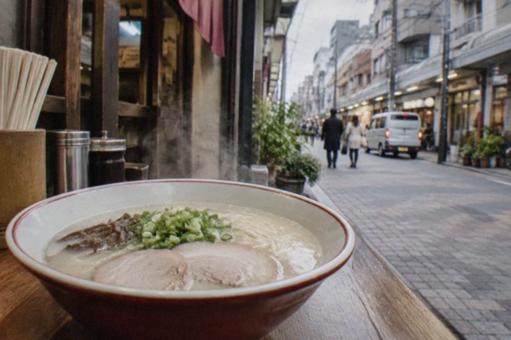 A steaming bowl of tonkotsu ramen with thin noodles and sliced pork on a wooden counter