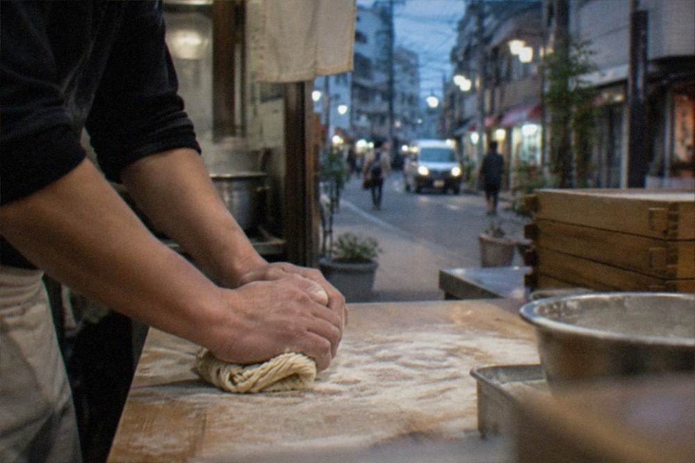 Hands kneading ramen noodle dough on a floured wooden board in a kitchen