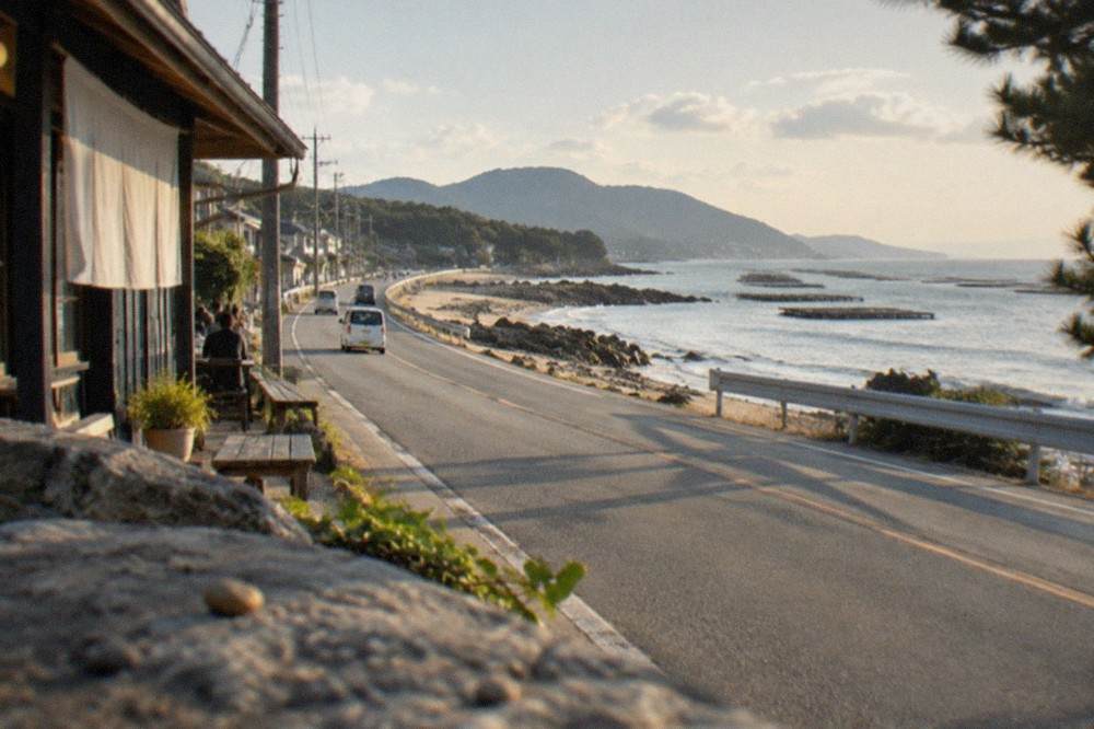 A long coastal road curving along a shoreline with oyster rafts visible on the water