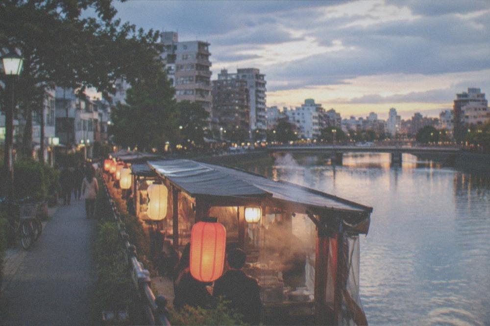Glowing food stalls lining a riverside street at night with lanterns and steam rising