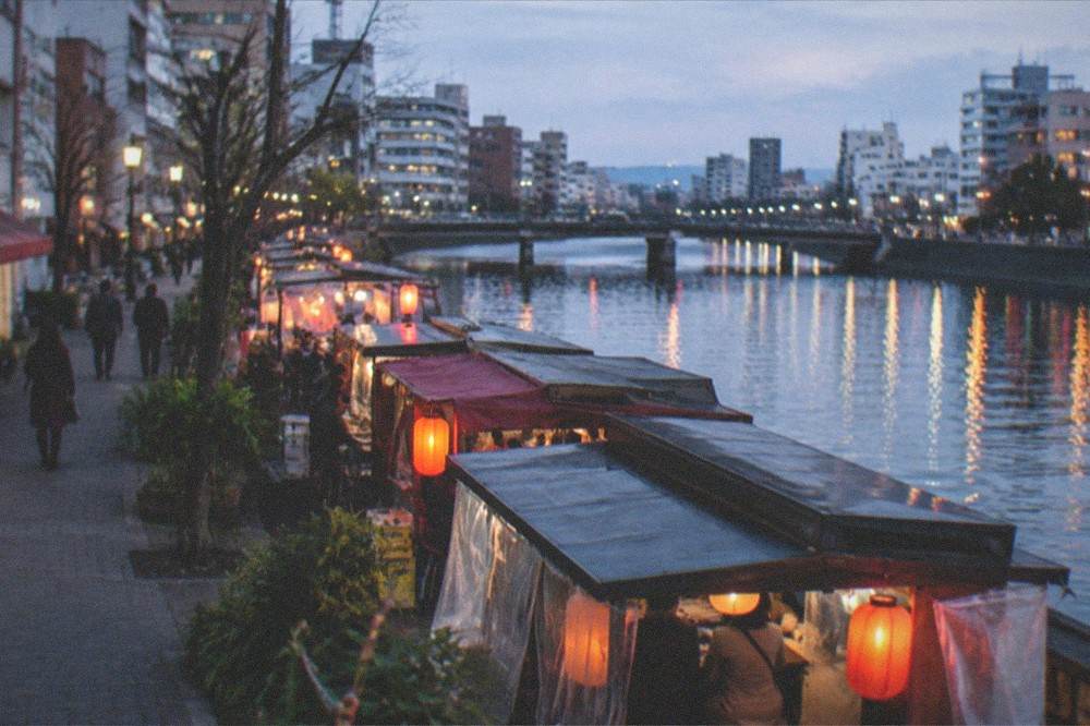Evening lights reflecting on a river beside a row of illuminated food stalls