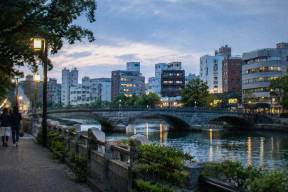 A park lake at sunset with a stone bridge and city skyline beyond