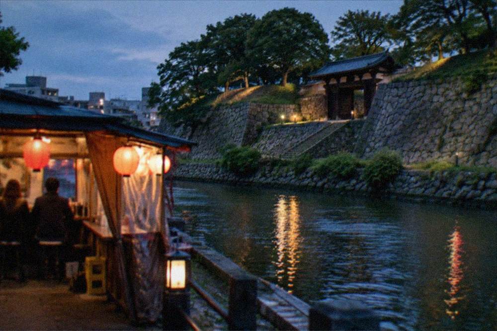 A lit food stall beside a river at dusk with lanterns reflecting on the water