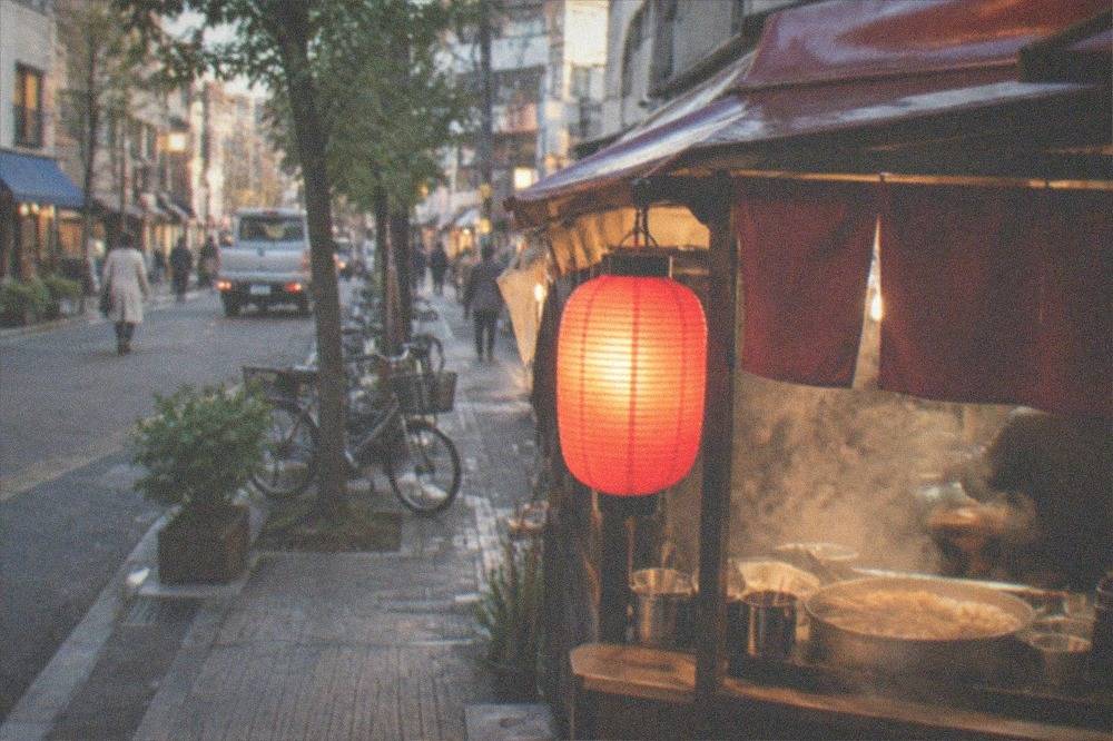 A food stall with a red lantern and steam rising from a counter at dusk