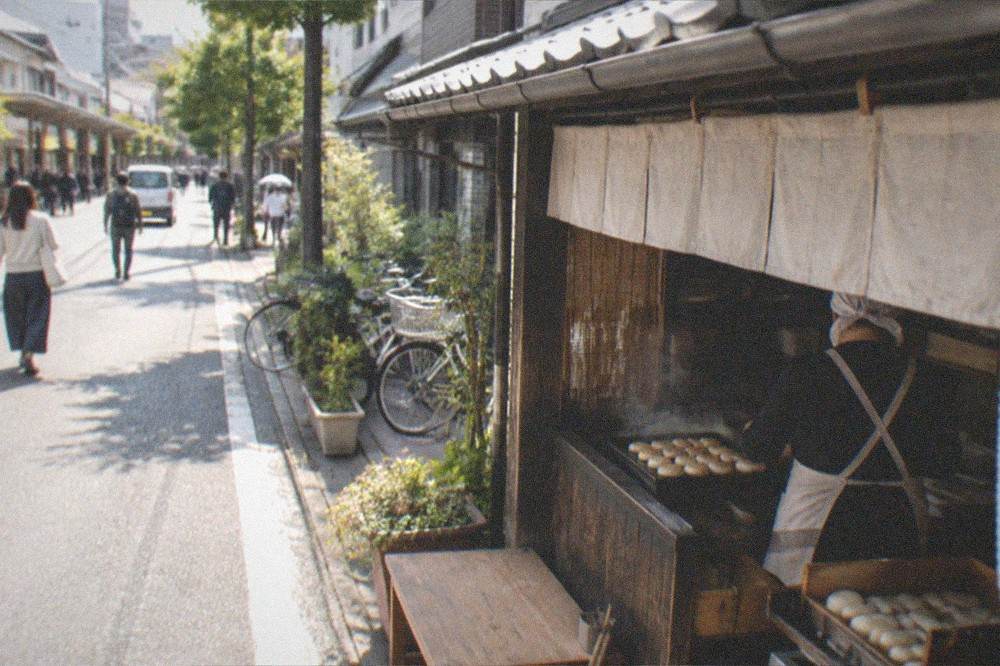 A shopfront grilling small rice cakes on a hot plate beside a historic street
