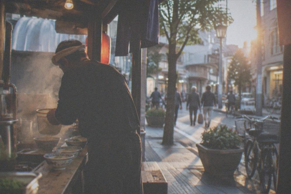 Chef preparing noodles at a small open-front food stall counter at dusk