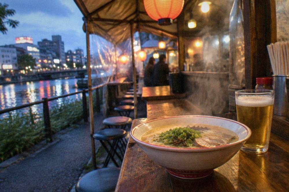 A steaming bowl of tonkotsu ramen on a wooden yatai counter beside a glass of beer
