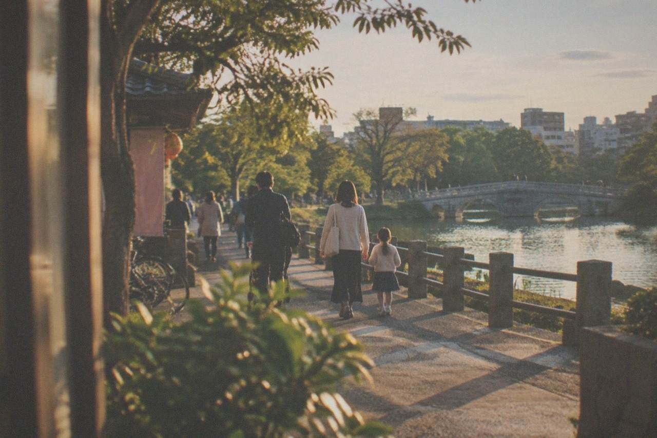 Families strolling beside a large pond with a stone bridge and green trees in a Japanese park